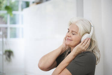 Relaxed senior woman enjoying music through headphones at apartment - JAHF00047