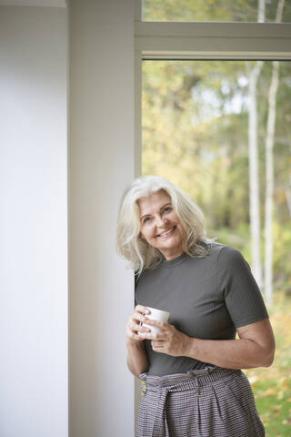 Lächelnde Frau im Ruhestand hält eine Tasse und lehnt sich an eine weiße Wand vor einem Fenster in einer Wohnung, lizenzfreies Stockfoto