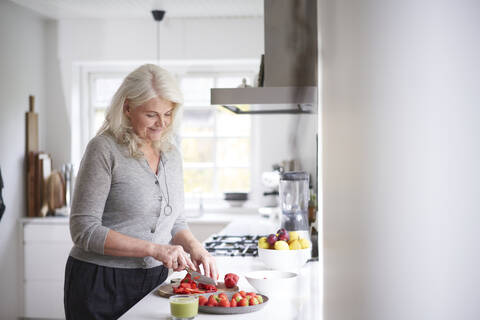 Frau im Ruhestand schneidet Erdbeeren auf einem Brett in der Küche zu Hause, lizenzfreies Stockfoto