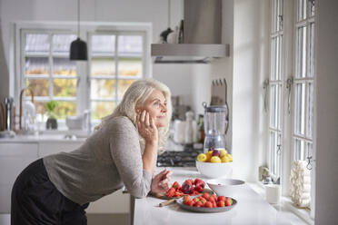 Nachdenkliche Rentnerin lehnt sich mit Erdbeeren in der Wohnung an den Küchentisch - JAHF00053