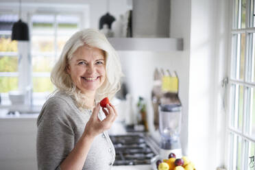 Lächelnde ältere Frau mit langen weißen Haaren, die eine frische Erdbeere in der Hand hält, während sie in der Küche zu Hause steht - JAHF00054