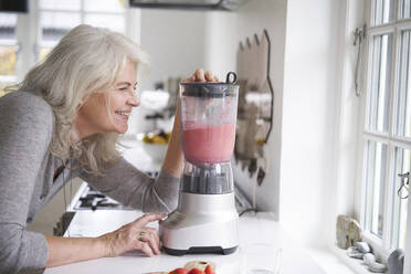 Lächelnde Rentnerin bei der Zubereitung eines Erdbeer-Smoothies in einer Küchenmaschine am Küchentisch - JAHF00057