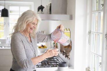 Fröhliche ältere Frau, die in der Küche einen Erdbeer-Smoothie in ein Glas gießt - JAHF00058