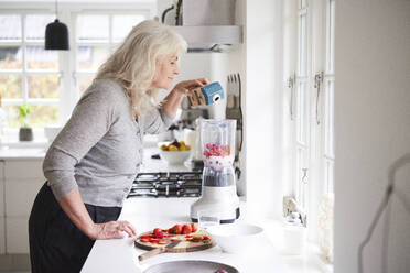 Ältere Frau gießt Milch in eine Küchenmaschine, während sie in der Küche einen Erdbeer-Smoothie zubereitet - JAHF00060