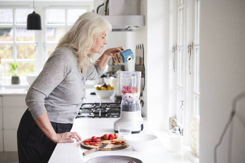 Ältere Frau gießt Milch in eine Küchenmaschine, während sie in der Küche einen Erdbeer-Smoothie zubereitet - JAHF00060