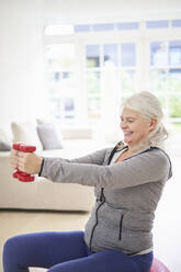 Lächelnde ältere Frau im Ruhestand, die zu Hause mit Hanteln trainiert - JAHF00064