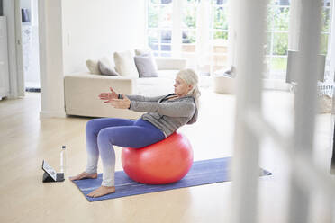 Ältere Frau, die auf einem Fitnessball trainiert, während sie über ein digitales Tablet zu Hause von einem Online-Tutorial lernt - JAHF00065