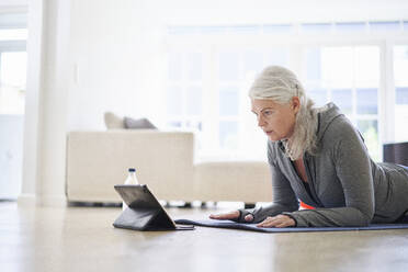 Eine ältere Frau im Ruhestand schaut auf ein digitales Tablet, während sie in ihrer Wohnung E-Learning-Übungen auf einem digitalen Tablet durchführt - JAHF00067