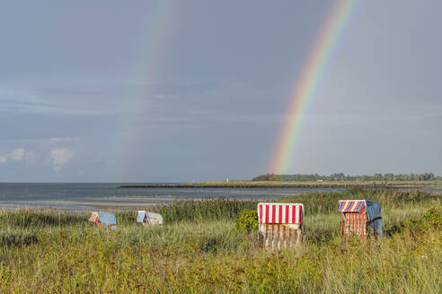 Doppelter Regenbogen, der sich über Strandkörbe mit Kapuze wölbt, die am grasbewachsenen Ufer stehen - KEBF01703