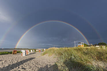 Double rainbow arching over hooded beach chairs standing on sandy beach - KEBF01704