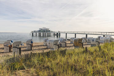 Deutschland, Schleswig-Holstein, Timmendorfer Strand, Strandkörbe mit Kapuze am sandigen Küstenstrand mit Teehaus am Ende der Seebrücke im Hintergrund - KEBF01723