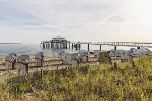 Deutschland, Schleswig-Holstein, Timmendorfer Strand, Strandkörbe mit Kapuze am sandigen Küstenstrand mit Teehaus am Ende der Seebrücke im Hintergrund - KEBF01723