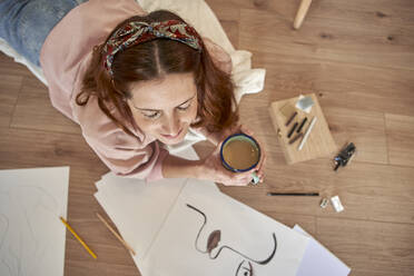 Female artist with drawings holding coffee cup while lying on floor at home - VEGF03354