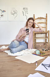 Lächelnde Künstlerin, die ein Mobiltelefon benutzt, während sie sich zu Hause an einen Stuhl lehnt - VEGF03366