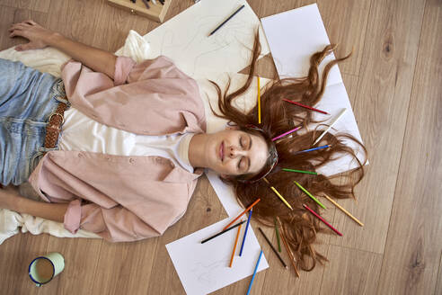 Smiling female artist lying down with colored pencil in hair while eyes closed at home - VEGF03371
