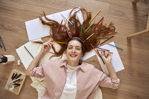 Smiling female artist with bunch of colored pencils in hair while lying down at home - VEGF03372