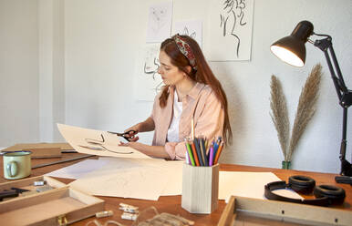 Female freelancer cutting paper edges at desk while working at home - VEGF03378