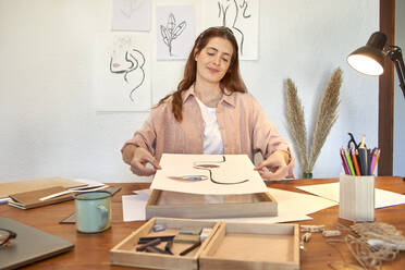 Female artist holding drawing over picture frame at desk in living room - VEGF03379