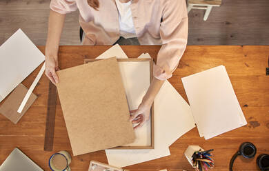 Künstlerin hält hölzernen Bilderrahmen am Schreibtisch, während sie zu Hause arbeitet - VEGF03381