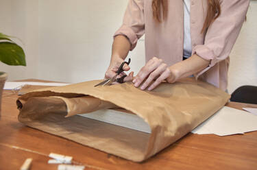 Künstlerin schneidet braunes Papier, während sie am Schreibtisch im Wohnzimmer ein Paket vorbereitet - VEGF03387