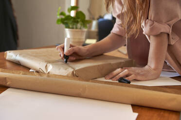 Female freelancer writing message on wrapped package at desk in living room - VEGF03390