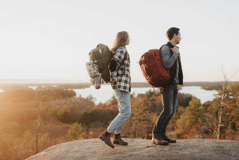 Junges Paar steht am Rande eines Felsvorsprungs und bewundert den umliegenden Wald bei Sonnenuntergang im Herbst - SMSF00515