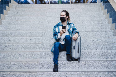 Man wearing protective face mask using mobile phone while sitting with luggage on steps - EBBF01899