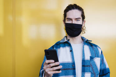 Junger Mann mit Schutzmaske und Mobiltelefon, der gegen eine gelbe Wand starrt - EBBF01901