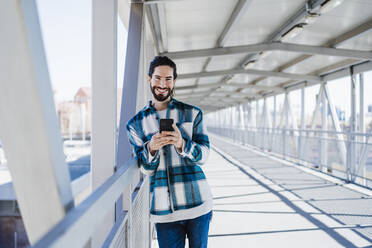 Lächelnder Mann, der ein Mobiltelefon benutzt, während er auf einer Brücke steht - EBBF01910