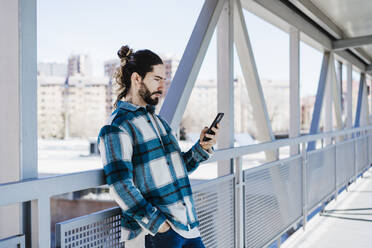 Young man using mobile phone while leaning on railing of bridge - EBBF01911