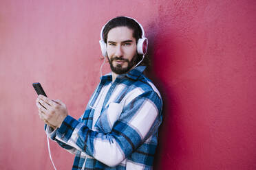 Junger Mann mit Kopfhörern, der ein Mobiltelefon benutzt und sich an eine rote Wand lehnt - EBBF01922