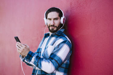 Young man wearing headphones using mobile phone while leaning on red wall - EBBF01922