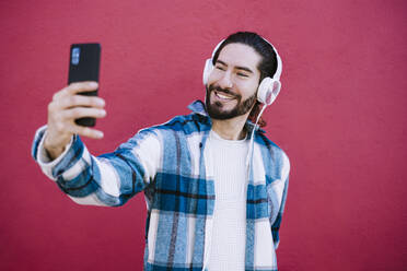 Smiling man wearing headphones taking selfie while standing against red wall - EBBF01923