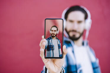 Man taking photo through mobile phone while standing against red wall - EBBF01924