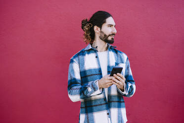 Young man with mobile phone looking away while standing against red wall - EBBF01925