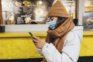 Junge Frau mit Smartphone vor einem Café im Winter während COVID-19 - OYF00292