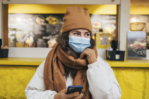 Junge Frau mit Hand am Kinn träumt, während sie ihr Smartphone in einem Café während der COVID-19 hält, lizenzfreies Stockfoto
