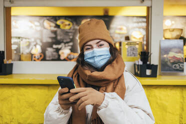 Junge Frau mit Gesichtsmaske und Mobiltelefon im Herbst in einem Café - OYF00294