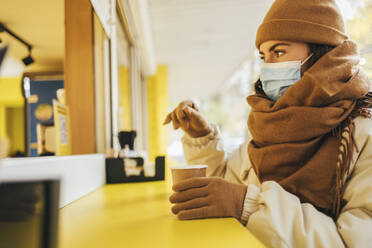 Junge Frau mit Maske und Einweg-Kaffeebecher in einem Straßencafé im Herbst - OYF00296