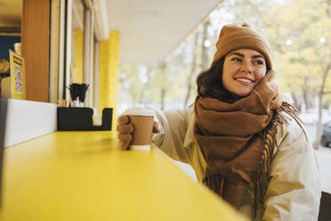 Lächelnde junge Frau mit Einweg-Kaffeetasse, die in einem Straßencafé im Winter wegschaut - OYF00297