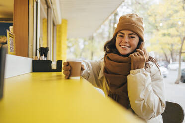 Happy young woman with disposable coffee cup at street cafe during autumn - OYF00298