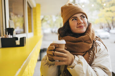 Smiling woman with disposable coffee cup day dreaming at street cafe during winter - OYF00299