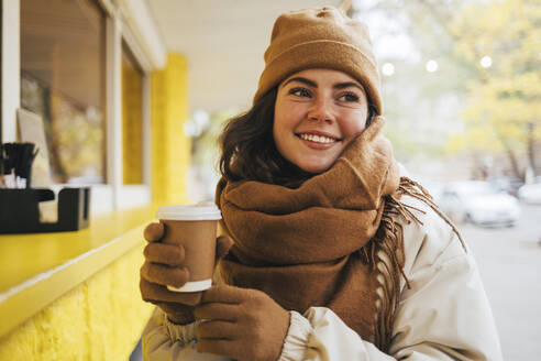 Lächelnde junge Frau, die mit einer Einweg-Kaffeetasse in einem Straßencafé im Winter wegschaut - OYF00301
