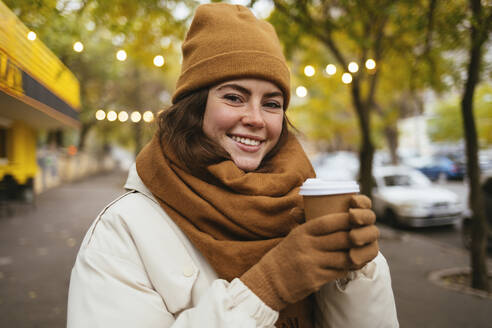 Glückliche junge Frau mit Strickmütze und Einweg-Kaffeetasse im Winter - OYF00304