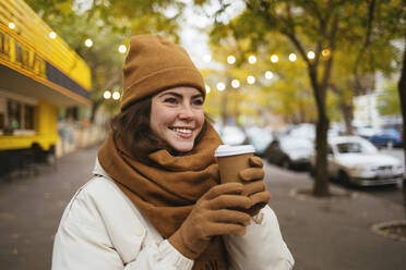 Happy woman wearing knit hat day dreaming with disposable coffee cup on street during winter - OYF00307
