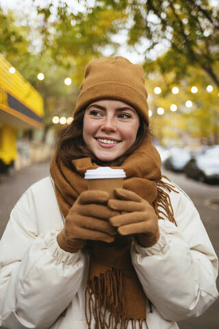 Lächelnde Frau in warmer Kleidung hält Einweg-Kaffeebecher auf dem Bürgersteig, lizenzfreies Stockfoto