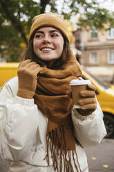 Happy young in brown scarf with disposable coffee cup on street during autumn - OYF00312
