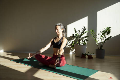 Yogini meditiert auf einer Übungsmatte im Wohnzimmer an einem sonnigen Tag - MCVF00666