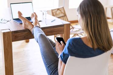 Brünette Geschäftsfrau, die ihr Smartphone benutzt, während sie sich mit hochgelegten Füßen am Tisch im Heimbüro entspannt - SBOF02286