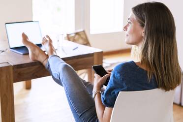 Lächelnde brünette Geschäftsfrau, die wegschaut, während sie mit Smartphone und hochgelegten Füßen am Tisch im Heimbüro sitzt - SBOF02287